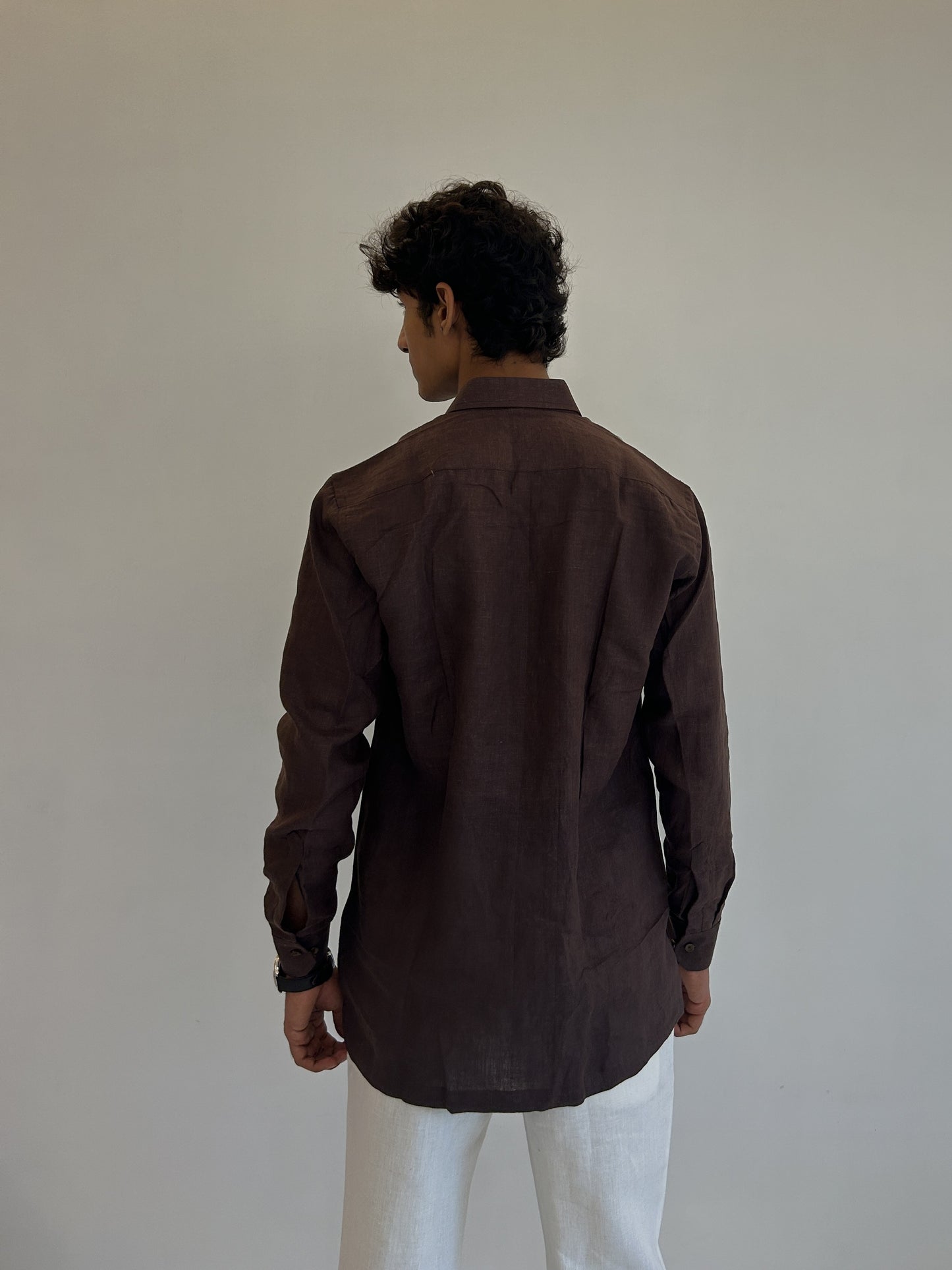 Back view of man wearing a chocolate brown linen shirt and white pants against a plain background