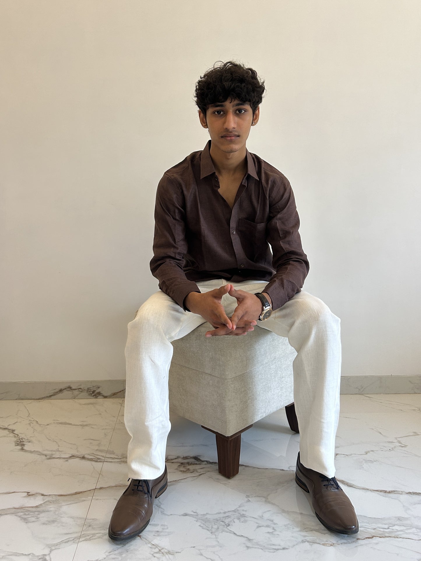 Man sitting on a chair wearing a brown linen shirt and white pants in a minimalistic room.