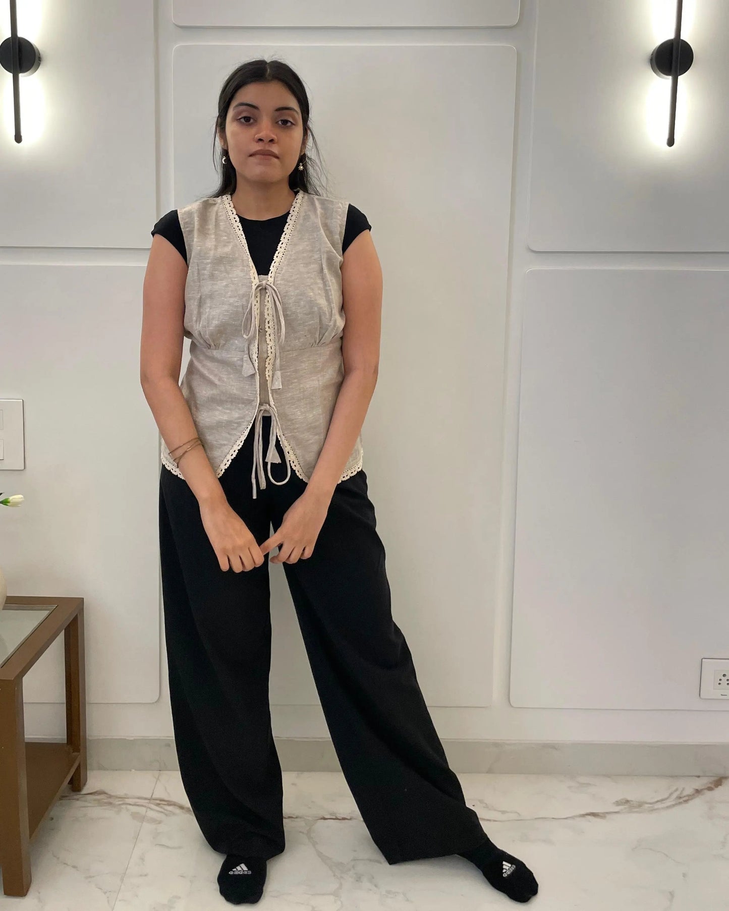 Person wearing a light-colored linen waistcoat over a black top and pants in a room with white walls and a wooden side table.