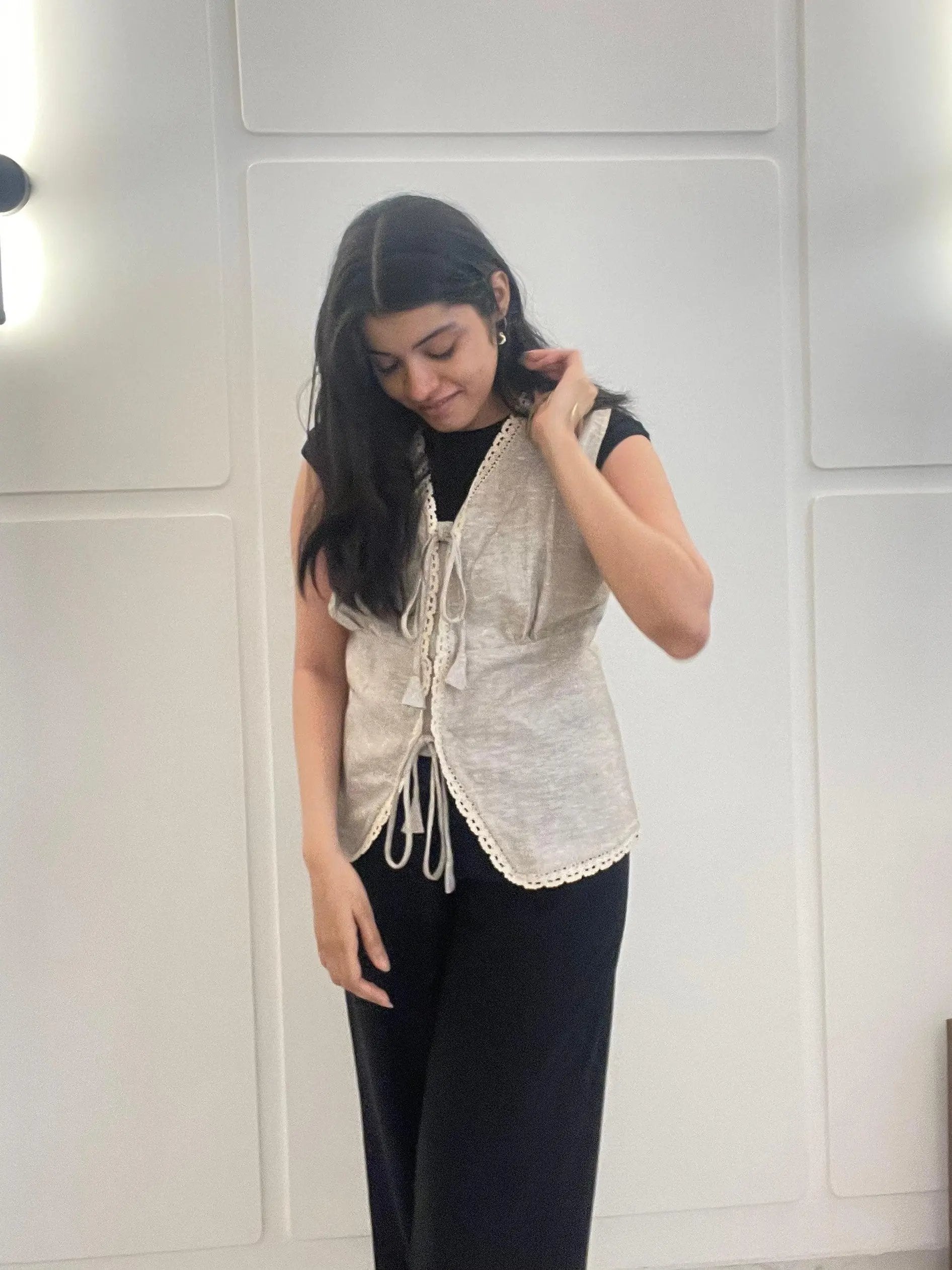A woman wearing a beige lace linen waistcoat over a black top and pants in front of a white wall.