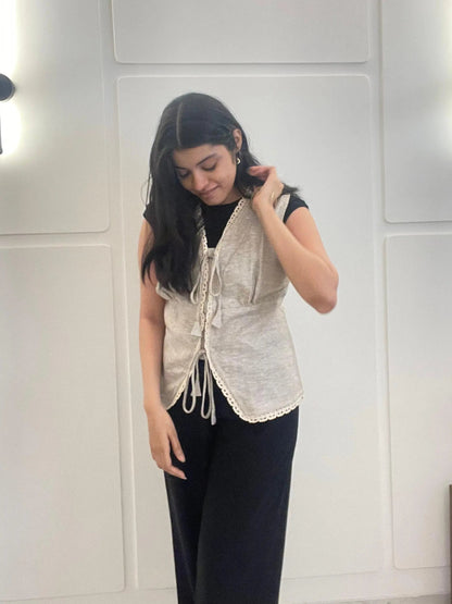 A woman wearing a beige lace linen waistcoat over a black top and pants in front of a white wall.